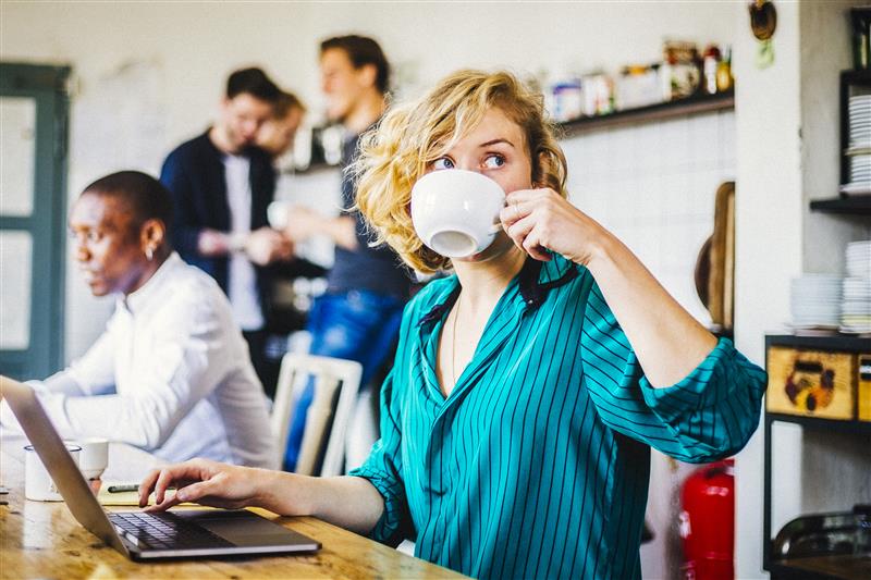A young woman sips her coffee and sits at a table with her laptop