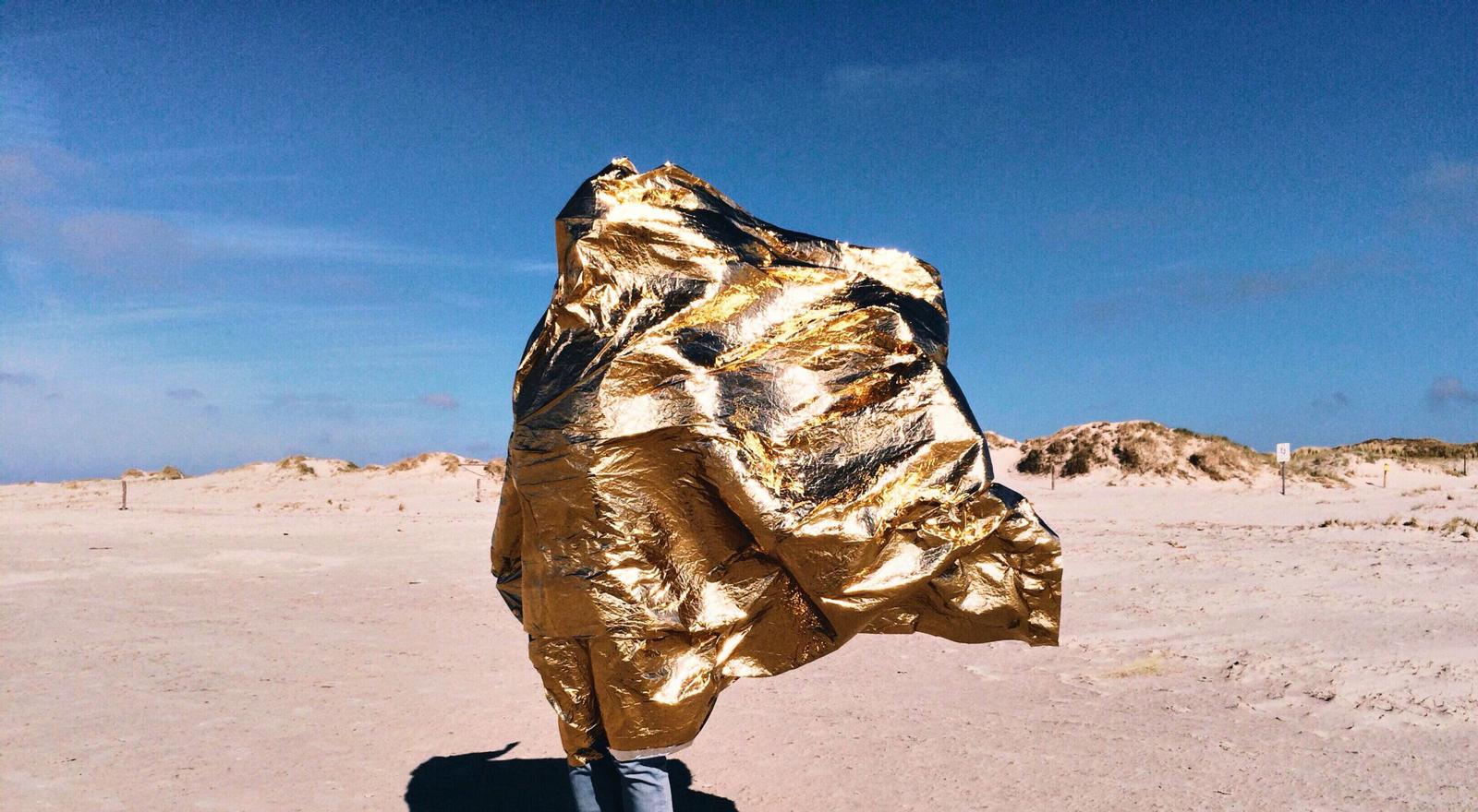Large Web and Screen A golden thermal emergency blanket is blown by the wind and covers a person standing on the beach