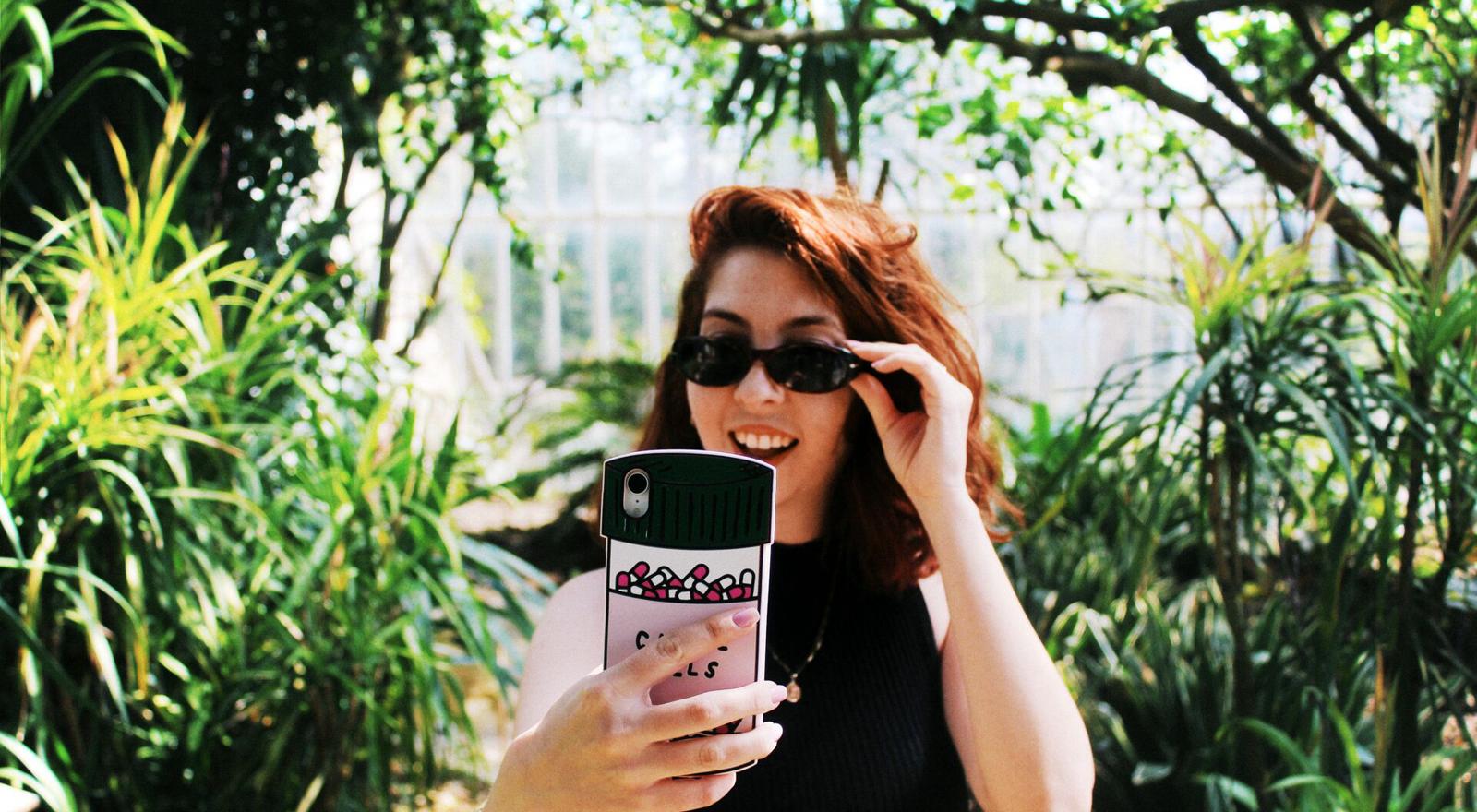 Large Web and Screen A young woman wearing sunglasses takes a selfie with her phone outside while surrounded by plants