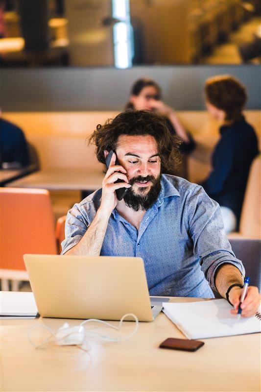Man with a beard taking notes while he's on the phone