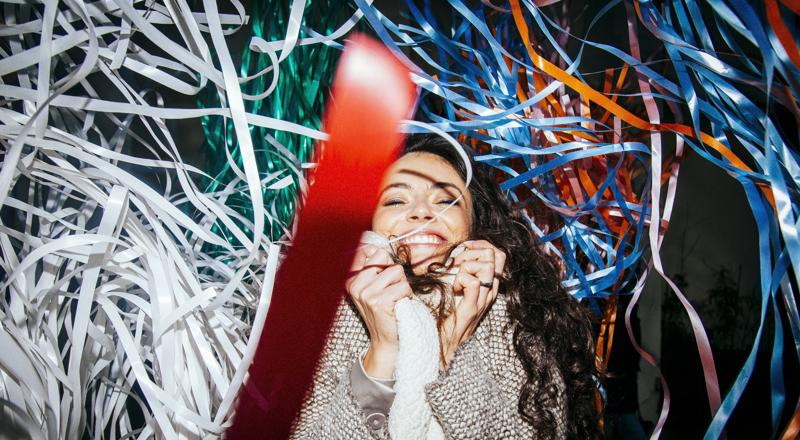 Young woman with long hair wraps coat around herself and smiles in the middle of some paper streamers